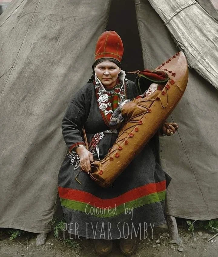 Outside the doorway of a lavvu — the traditional Sámi tent — a mother sits grounded and strong, holding her child in one of the most iconic carriers of northern Europe: the komse, or Sámi cradleboard.
This cradleboard, made from a single carved piec