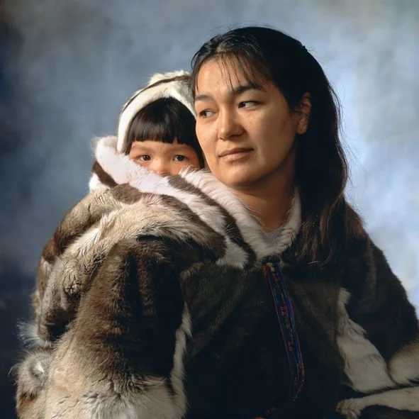 Wrapped in rich, fur-lined warmth, a mother from the Inuit community carries her child nestled deep inside the back of her traditional parka — known as an amauti. The amauti is more than winter clothing; it is a protective cocoon designed specificall