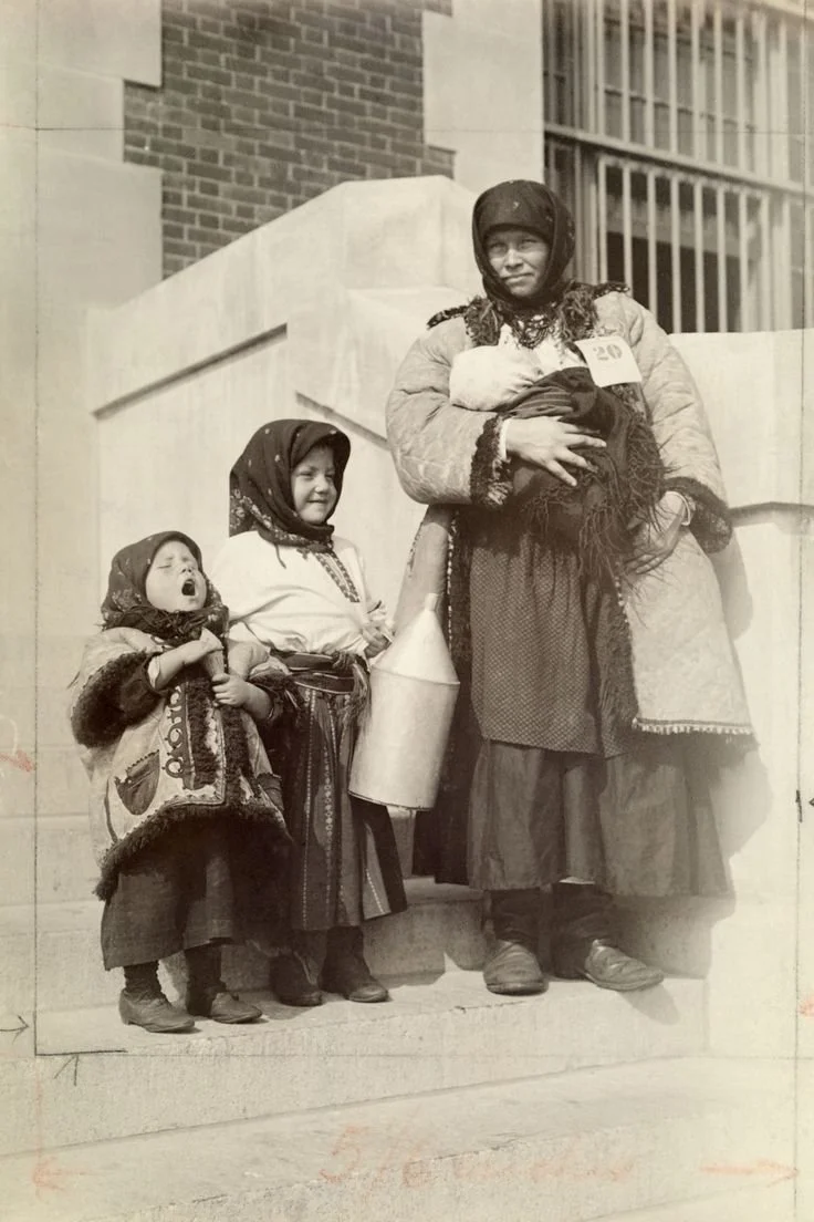 A mother stands with her three children on the stone steps of what appears to be an institutional building — most likely Ellis Island in New York, judging by the numbered tag pinned to her coat. She holds her swaddled baby close against her chest, wr