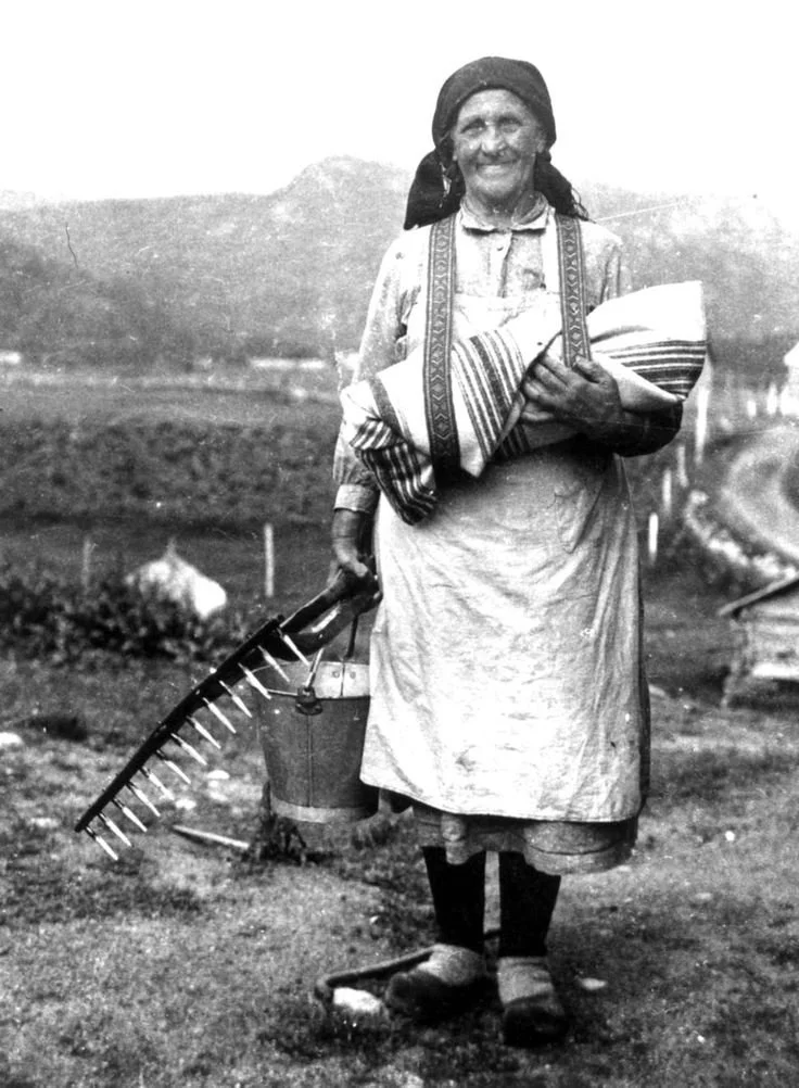 A woman stands smiling on the open land, her face weathered with age and sun, her hands full of the day’s tasks. In one arm she cradles a tightly swaddled infant, secured in woven fabric and bound close to her chest with wide straps that cross over h