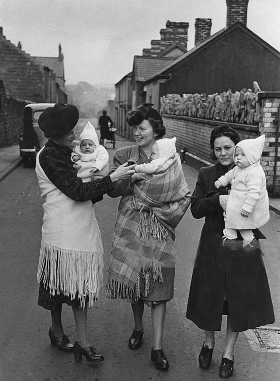 Three women walk side by side down a narrow street in postwar Wales, each carrying a baby wrapped against the chill. The woman in the centre stands out — her baby is tucked securely into a checked woollen shawl, worn in the traditional Welsh shawl ca