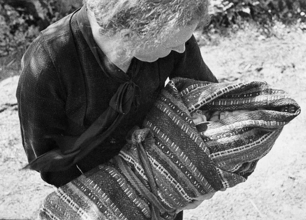 Cradled with devotion and wrapped in a richly textured blanket, this baby rests within a linde — the traditional infant carrier used in southern Norway. The woman, dressed in dark, formal attire and with her hair curled in careful waves, gazes down a