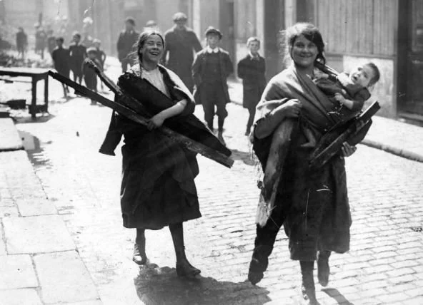 Two women walk arm-in-arm down a cobbled street, each carrying firewood, their laughter suspended in motion. One of them balances a young child on her hip, held close with a long shawl wrapped diagonally across her chest. The child’s legs hang freely