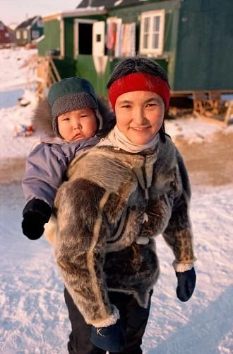 Standing on a snowy path in a northern village, a Greenlandic Inuit mother smiles warmly as she carries her bundled child high on her back in the traditional way. The child, well-dressed for the cold in a fur-lined parka and mittens, is nestled into
