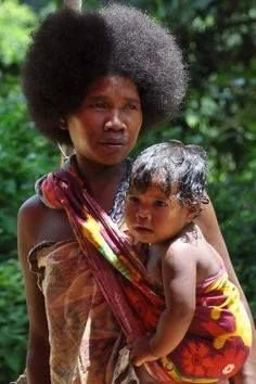 A mother gazes into the distance, standing tall and strong, with her child cradled closely in a vibrant floral wrap. The baby is carried on her front in a traditional torso-style sling made from colourful cloth, knotted over one shoulder. The wrap su