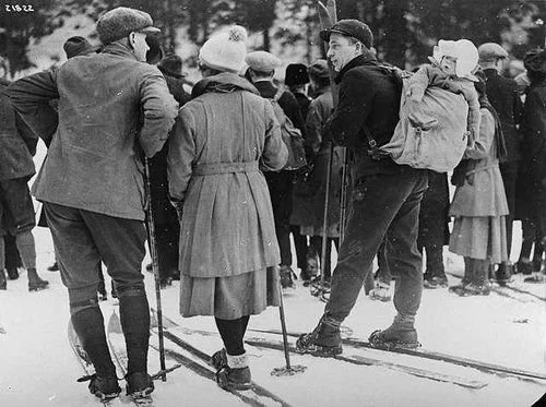 This striking historical photograph captures a group of Nordic skiers gathered in the snow, dressed in heavy wool clothing and caps typical of the early 20th century. Prominently featured is a man carrying a small child on his back using a structured