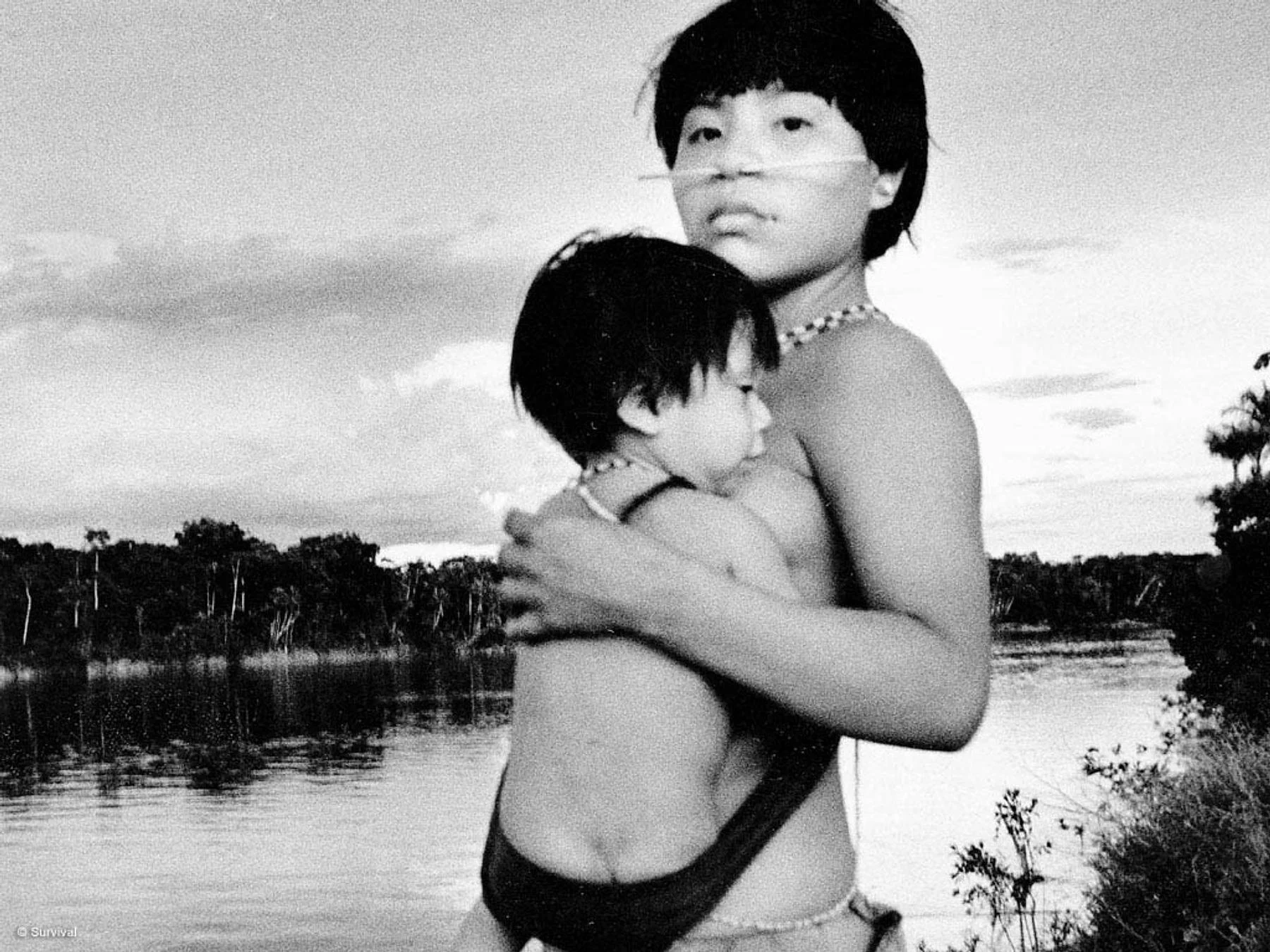 By the edge of the river, a young mother holds her sleeping child close, his little body wrapped snugly in a woven sling. Her gaze meets the camera with quiet strength, her face painted with fine white lines that mark identity and belonging. The wate