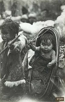 In this poignant historical photograph, a young child sits nestled in a traditional cradleboard — most likely from an Indigenous community of Siberia or the Russian Far East. The cradleboard’s oval frame, padded interior, and curved protective arch c