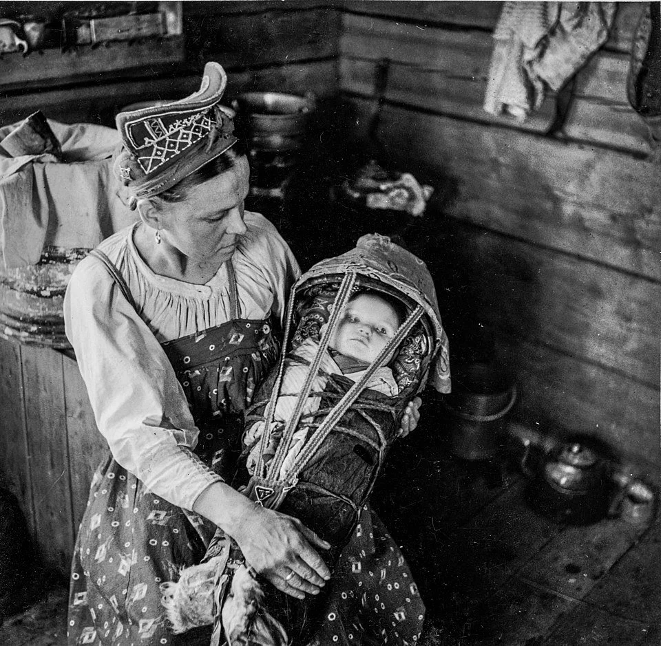 Inside a wooden cabin in the far north, a Skolt Sámi mother cradles her baby in a beautifully constructed cradleboard — known as a komsio in Northern Sámi. The baby lies snugly swaddled within the wooden frame, cushioned by layers of soft padding and