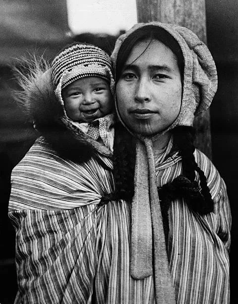 In this striking historical portrait, a mother from the Arctic region carries her child high on her back, wrapped snugly in a thick, striped textile. The child peeks over her shoulder, face framed warmly in fur, eyes wide and watchful against the icy