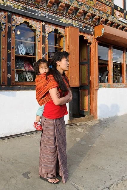 A young woman stands calmly outside a traditional shopfront, the colourful timberwork and hand-painted motifs behind her unmistakably Bhutanese. Her stance is relaxed but attentive, one arm across her chest and the other supporting the secure knot of