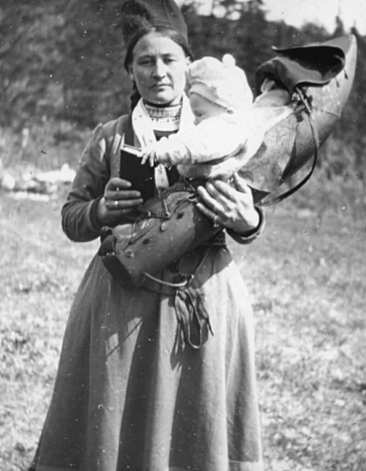 Wearing the distinctive four-cornered hat of a Kola Sámi woman, this mother holds her baby cradled in a traditional Sámi komsá or cradleboard. Photographed in the Russian Far North, her embroidered collar, belt tassels, and long pleated skirt reflect