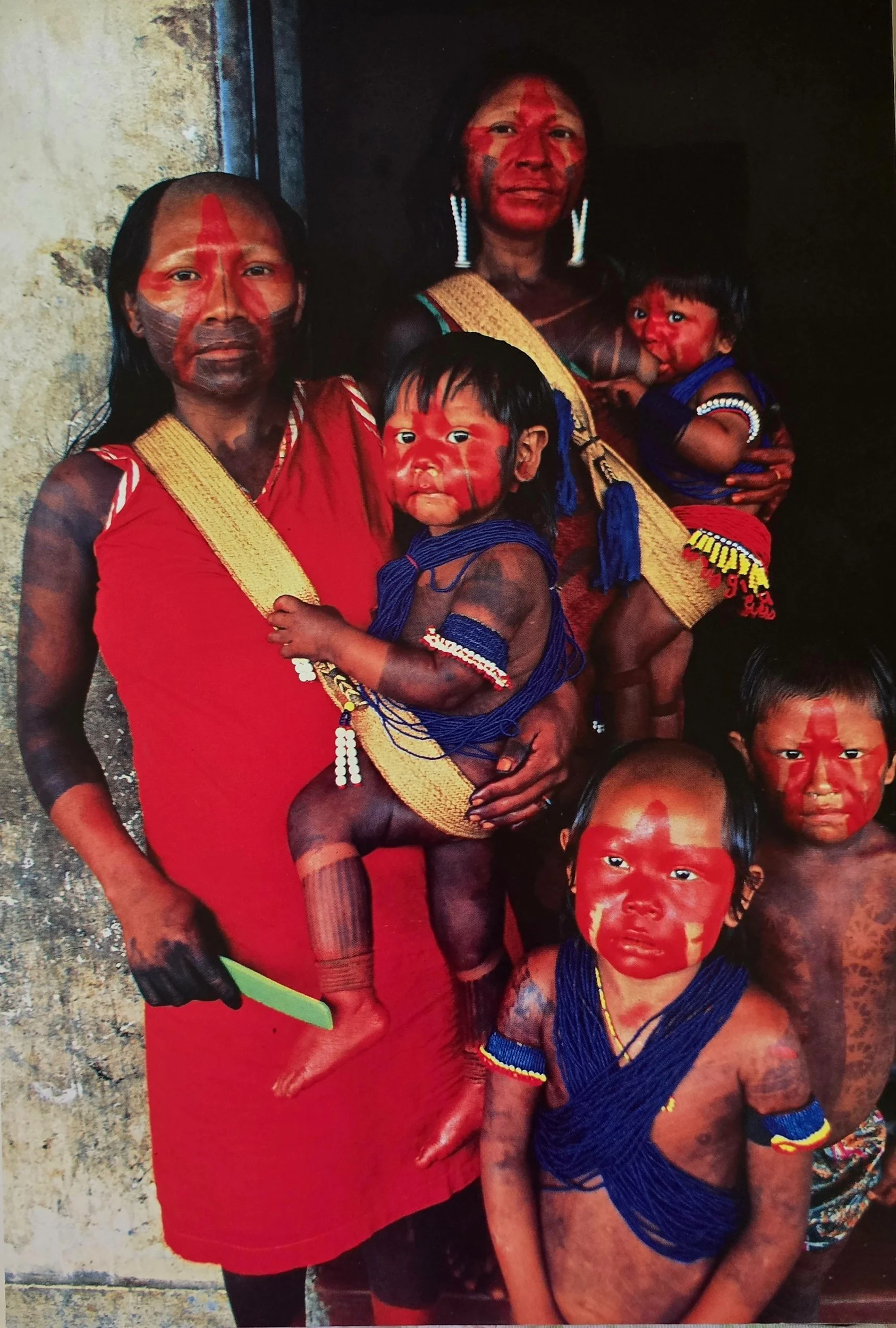 At the threshold of a concrete dwelling, a Kayapo family gathers in powerful stillness, their presence both intimate and defiant. Two mothers stand side by side, each carrying a young child in a handwoven sling worn across the chest—a deeply familiar
