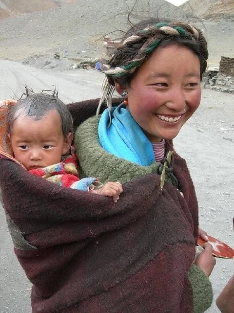 Wrapped snugly against the cold, a mother carries her baby high on her back in a thick woollen blanket. The baby is bundled deep into the carrier, just their head and hands visible beneath warm layers. The mother’s hair is braided and adorned with tr