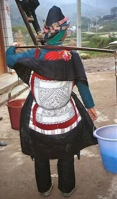 A Hmong woman walks with purpose along a village path, balancing a yoke across her shoulders, a red bucket on one end and a blue one on the other. But it’s not just water or supplies she’s carrying — on her back, nestled snugly within a beautifully e