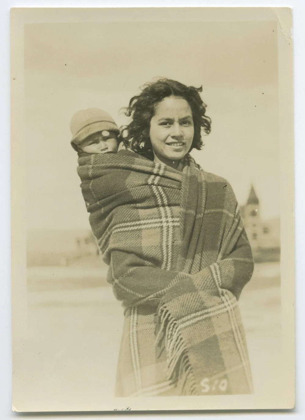 A young Māori mother stands in the open air, her smile warm and steady despite the chill hinted at by the tightly wrapped woollen blanket that holds her baby close. The child’s knitted cap and peeking eyes suggest the sharpness of the breeze, but nes