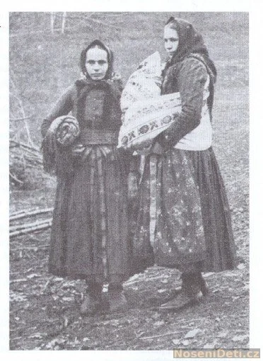 Two women stand together on a bare hillside, their traditional layered skirts brushing the cold earth. Both wear patterned aprons and headscarves, with thick, dark outer vests fastened tightly against the weather. One woman holds a large bundle, wrap