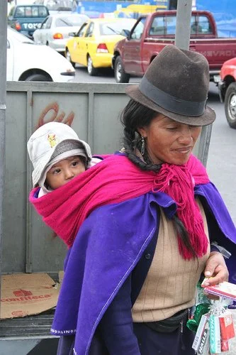 A woman in the Ecuadorian highlands carries her child close on a busy city street, wrapped snugly in a brightly coloured shawl. Her distinctive felt hat, thick braids, and layers of warm wool mark her as part of one of the many Kichwa-speaking Indige