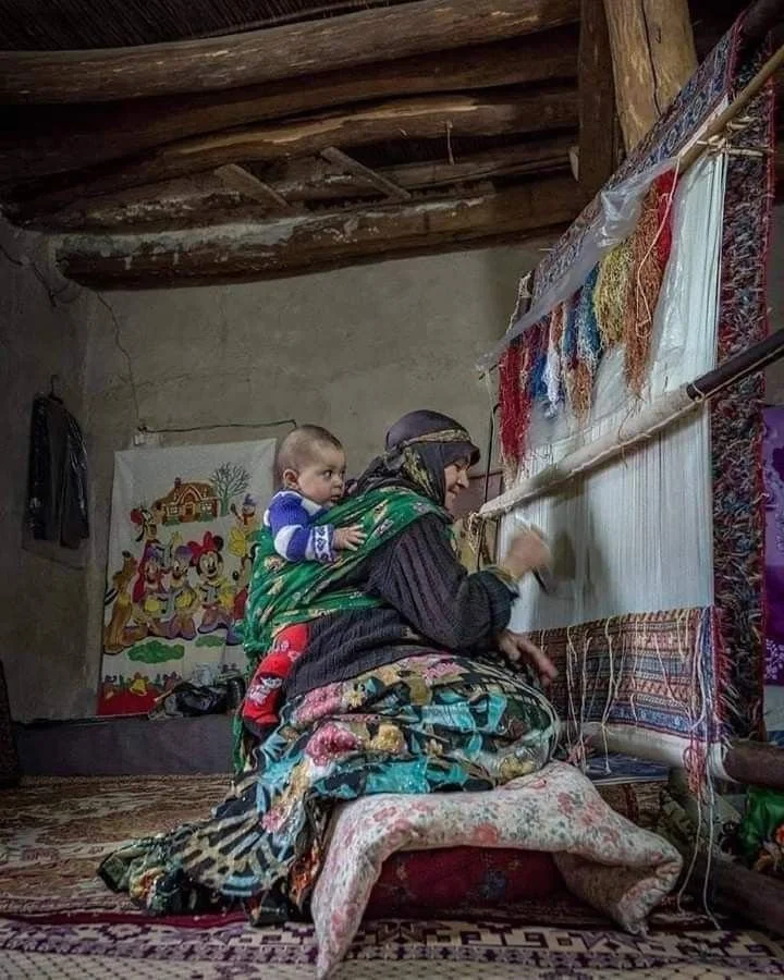 In this vivid image, a woman sits at a traditional upright loom, her hands expertly knotting a carpet as threads of many colours hang above. Behind her, wrapped snugly in layers of cloth and secured across her back with a long woven shawl, rests a yo