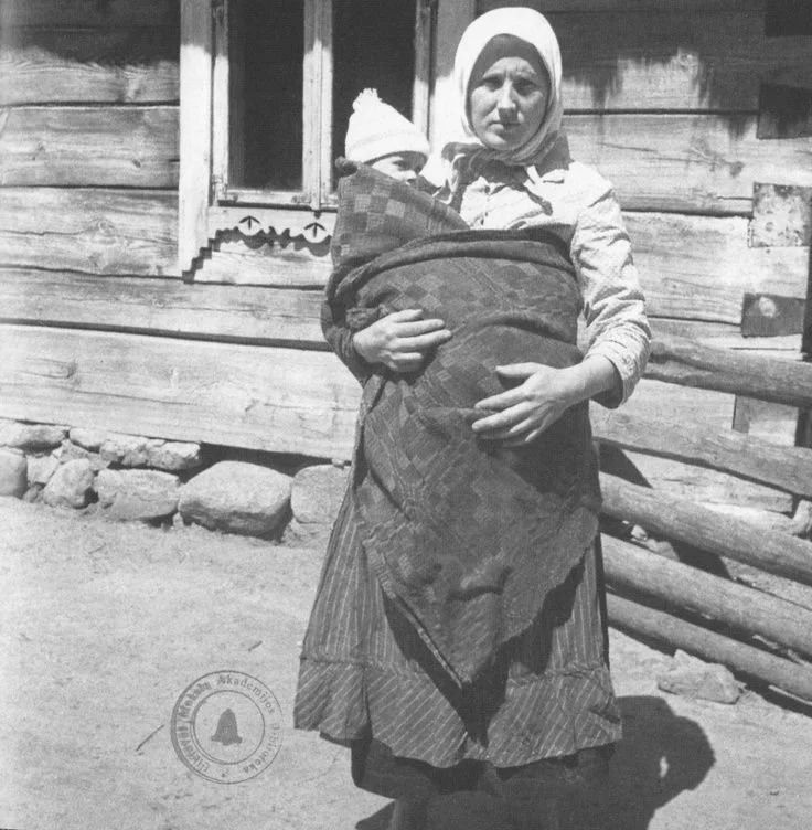 Outside a timber house in the Carpathian foothills, a mother stands quietly with her child wrapped against her chest. Her face is serious, her stance grounded, the lines of daily life etched in sun and shadow.
The woven wrap crosses diagonally acros