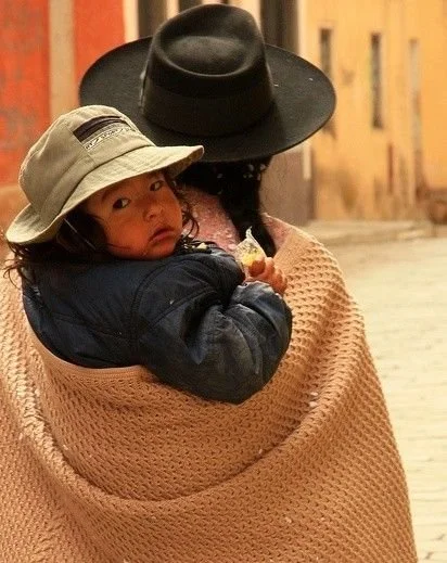 A mother walks down a narrow street in an Andean town, her dark braid tucked beneath a wide-brimmed black hat, her shoulders wrapped in a thick woollen manta. The woven shawl, a staple of daily life in the highlands of Bolivia and southern Peru, is g