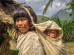 A mother from the foothills of Colombia’s Sierra Nevada smiles as she turns toward the camera, her child nestled high on her back in a soft cloth sling. The carrier—simple, secure, and beautifully adapted to the region’s terrain—is formed from a sing