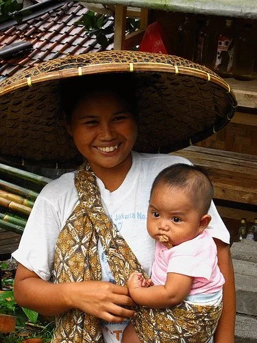 A smiling mother holds her baby snugly in a batik sling, wrapped across one shoulder and under the opposite arm in a traditional hip carry. The cloth is knotted high on her chest, allowing the patterned fabric to support the baby securely while leavi
