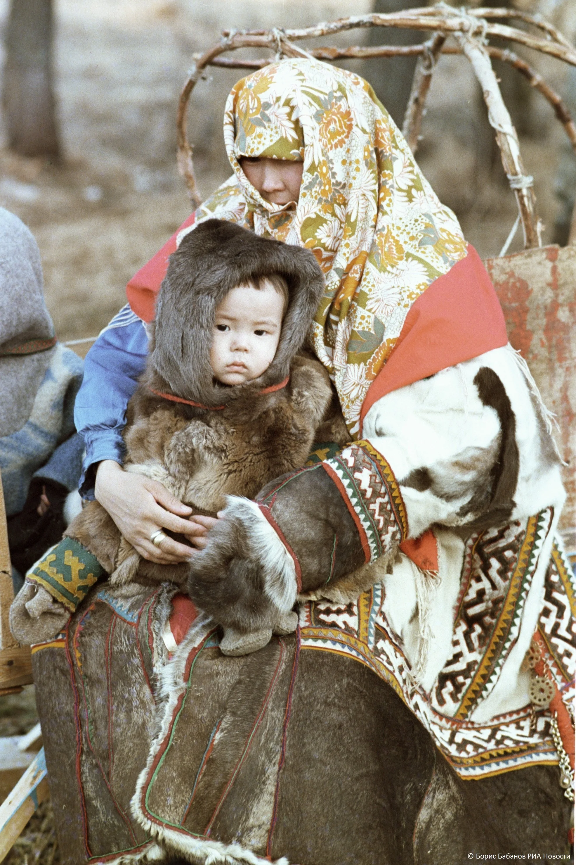 Wrapped in thick furs and seated within a traditional sled frame, this mother from the Siberian taiga cradles her child in a garment that is both carrier and cocoon. The pair are dressed in elaborately decorated reindeer-hide clothing, with embroider