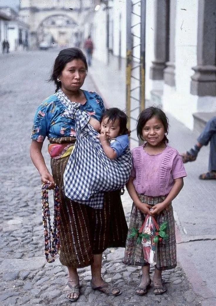 In this candid street portrait, a mother carries her toddler in a makeshift sling fashioned from a simple blue-and-white gingham cloth. The child is seated in a secure pouch formed by the tied ends, with the fabric looped over the mother’s shoulder a