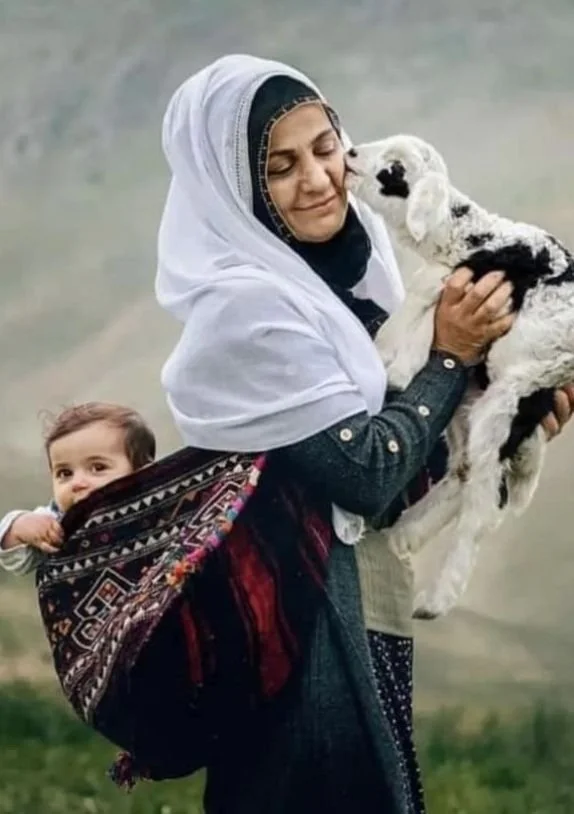 In a lush, mountainous landscape, a woman smiles gently as she holds a lamb close to her face, receiving a tender nuzzle. Draped in a long, dark dress and a white headscarf, she radiates a sense of calm and deep connection — not just with the lamb in
