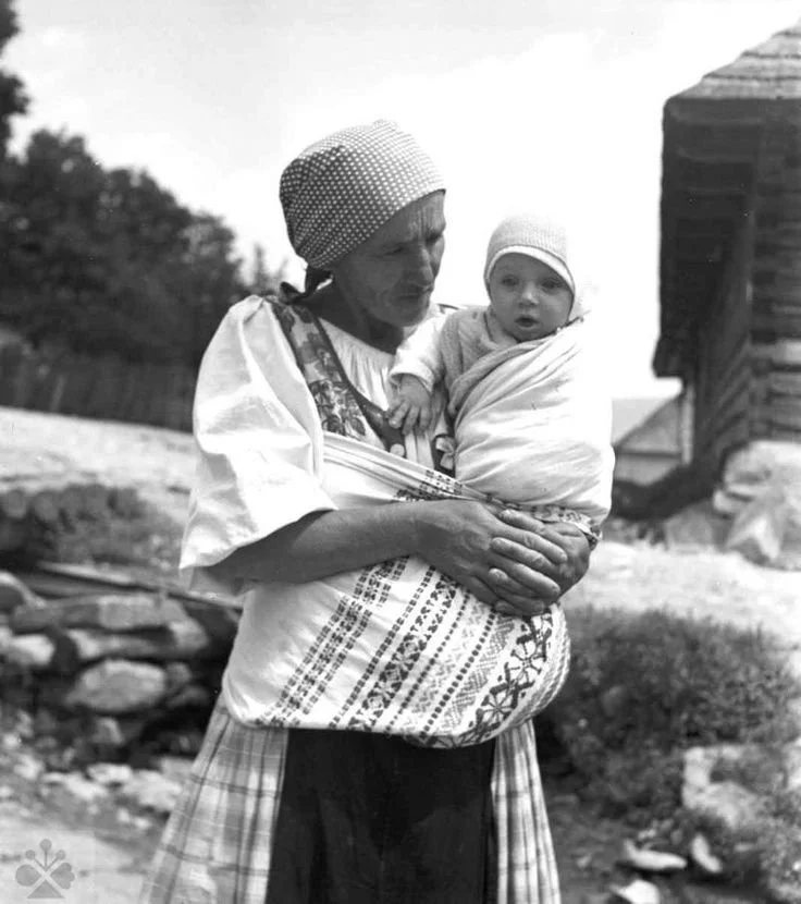 A mother stands outside a timber home in a rural village, her baby wrapped securely in a wide, embroidered cloth tied across her body. The woven sling rests high on her hip, holding the infant close in a seated position, one arm free, eyes wide and c