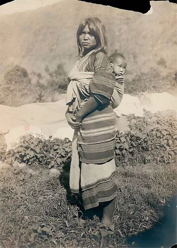 In the cool highlands of northern Luzon in the Philippines, a woman stands tall, her baby resting quietly against her back. Woven cloth wraps snugly around them both, anchoring the child in place with practiced ease. Her layered, striped garments and