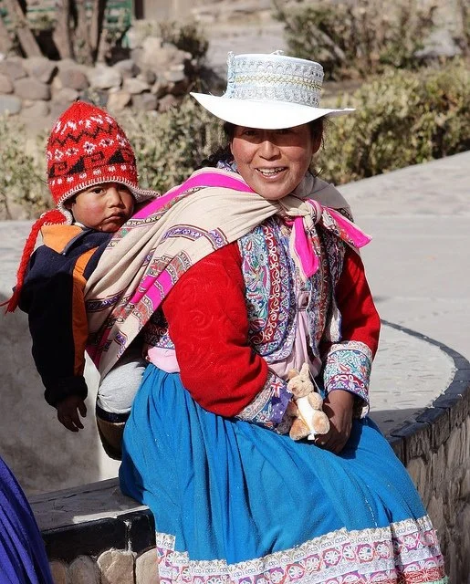 A mother in the Colca Valley of southern Peru sits resting on a stone bench, her warm smile framed by the crisp white brim of her traditional hat. Her clothing is richly embroidered in the distinctive style of the region—each flower and swirl stitche