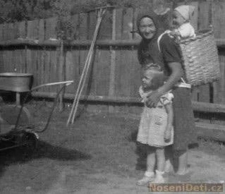 In this charming backyard moment, a smiling woman stands with one arm wrapped around a small child, while another baby rides high on her back in a large woven basket. The basket is strapped over her shoulders with what appear to be simple textile or