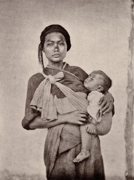 A quiet strength emanates from this sepia-toned portrait of a young mother, likely photographed in the late 19th or early 20th century. Standing against a bare wall, she gazes directly at the camera, her expression composed yet intense. Her baby slee