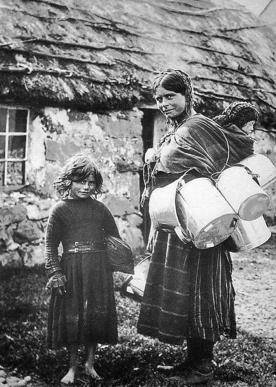 This evocative black-and-white photograph offers a rare and powerful glimpse into rural Irish life, most likely from the late 19th or early 20th century.
A barefoot girl stands beside her mother, who is carrying multiple tin pails—likely used for co