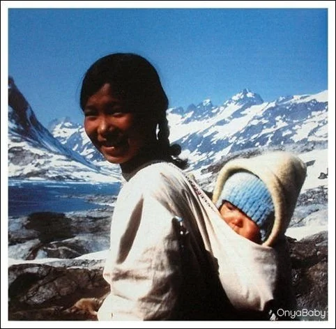 A Greenlandic Inuit mother smiles beneath a bright arctic sky, snow-capped peaks rising behind her. Her baby is carried snugly on her back in a warm cloth carrier, bundled in layers including a blue knitted hat and thick hood. The carrier is tied hig
