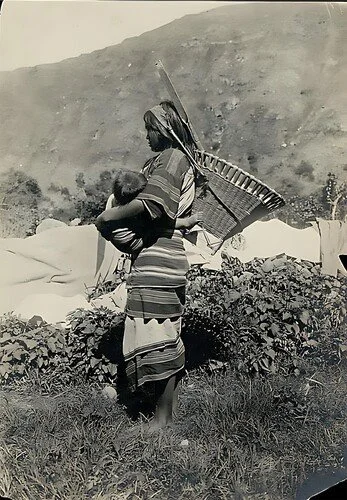 A woman of the Cordillera region in northern Luzon, Philippines, carries her baby securely in a woven cloth wrap across her front, close to her body. Her striped woven dress and the large basket carried with a tumpline over her head are typical of th