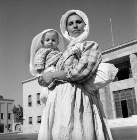 A mother stands tall in the sunlit square, her gaze steady and strong. Dressed in traditional rural clothing — a full skirt, checked blouse, and a headscarf knotted under her chin — she carries her baby in her arms, while a long cloth bundle is tied