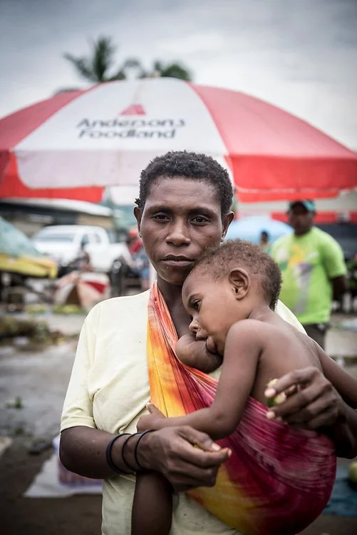 In this intimate portrait, a mother from Papua New Guinea holds her child close using a simple cloth sling draped over one shoulder. The bright, tie-dyed fabric supports her child snugly against her chest, allowing the little one to rest securely whi