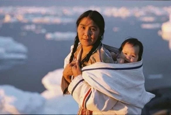 An Inuit woman carries her baby on her back, snugly wrapped in a traditional amauti — a thick, warm parka with an extended hood designed specifically for carrying young children. The child is held upright between the woman’s back and the garment’s br