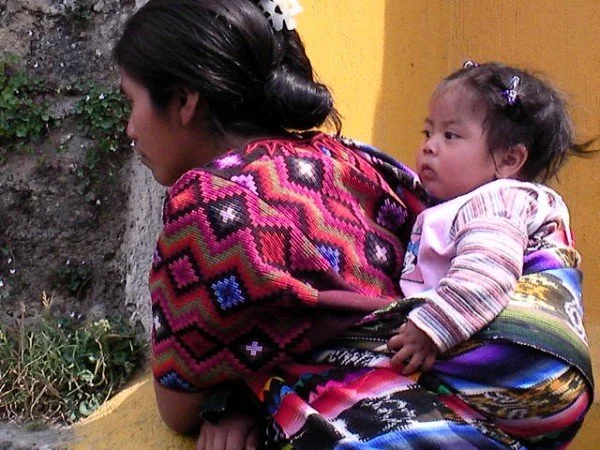 This beautiful image shows a Maya mother in Guatemala carrying her baby on her back in a traditional tzute. The bright, intricately woven cloth wraps the baby securely and keeps them close as she moves through her day. Each tzute carries unique patte