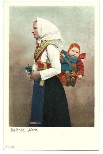 A mother from Mora in the Swedish region of Dalarna stands poised in full folk dress, her baby nestled high on her back in a textile carrier that blends seamlessly with the bold colours and patterns of local tradition. Both mother and child wear the