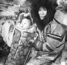 In this striking archival image, a mother from the Russian Far East cradles her baby in a traditional upright carrier, expertly crafted for warmth, safety, and mobility. The infant is securely nestled in a fur-lined cradleboard, wrapped against the c