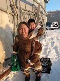 Bathed in golden Arctic light, an Elder carries a smiling child in the warm embrace of a traditional Inuit amauti. The child’s joyful face peeks over her shoulders, nestled snugly in the built-in pouch that sits between the shoulders and back — a des