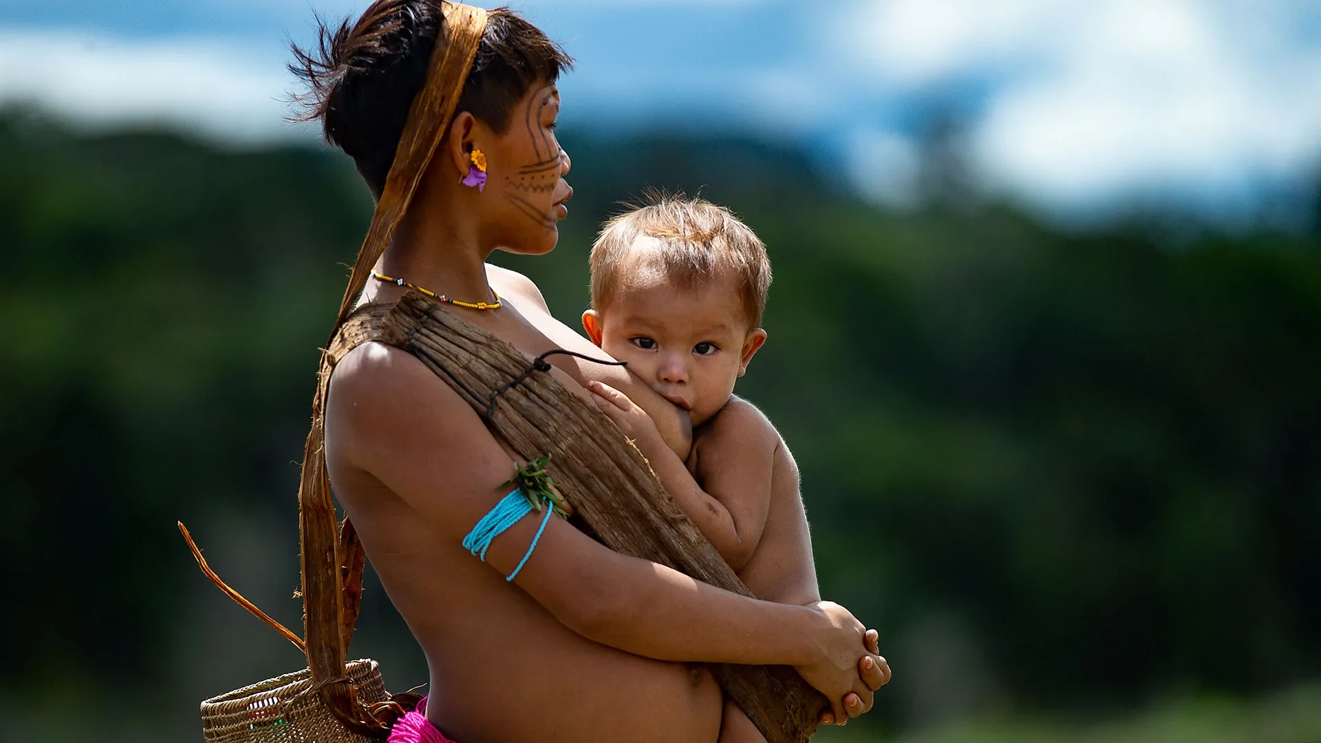 A mother from the Amazon rainforest cradles her child close as they share a quiet moment of breastfeeding. Her woven fibre headband and carrying band—likely crafted from natural palm or plant fibres—cross her chest diagonally, anchoring the child aga