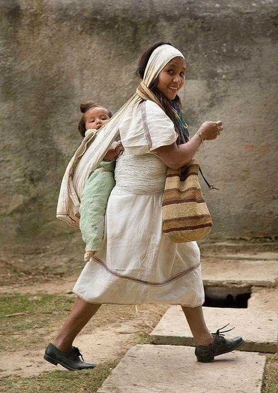 A mother walks with confident ease, wrapped in the daily rhythm of caregiving. Her child rides high on her back in a rebozo-style carrier, the cloth drawn snugly over one shoulder and tied beneath the opposite arm. This instinctive method is deeply r
