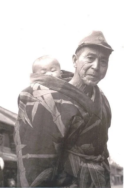 An elderly man stands in quiet strength, his face deeply lined with years of lived experience. Draped in a patterned kimono and cap slightly askew, he carries a sleeping baby on his back with calm assurance. The cloth—likely an extension of his own r