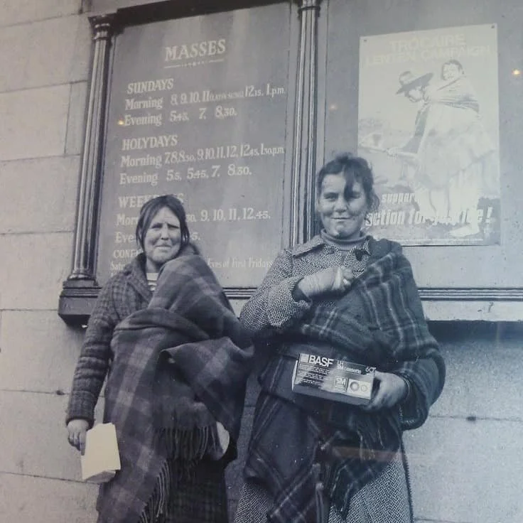 Two women stand outside a church noticeboard in Ireland, wrapped in large checked shawls with children held close. Behind them, a timetable for Masses is posted on the wall, and to the right, a Trócaire Lenten Campaign poster shows another mother in
