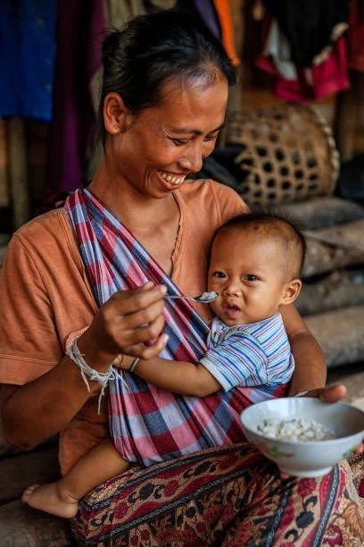 A mother in Laos shares a joyful moment with her baby, who sits nestled in a traditional checked pha khao ma cloth sling. As she offers a spoonful of rice porridge, both smile — the baby's expression a delightful mix of curiosity and mischief. The sl